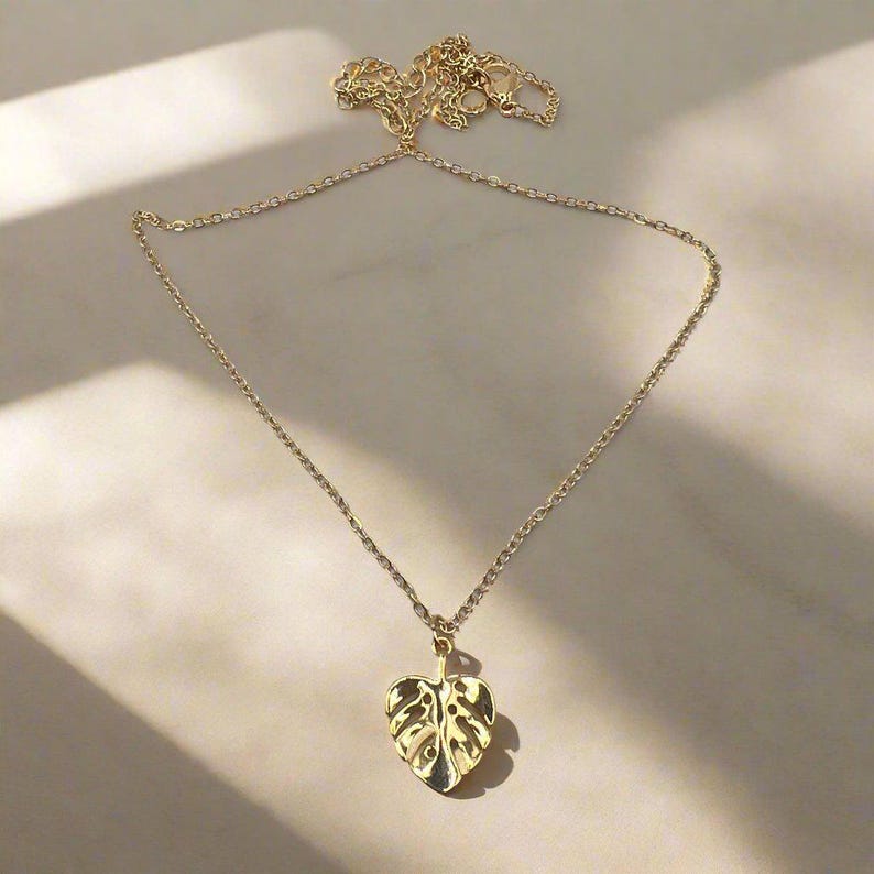 May include: A gold chain necklace with a delicate gold monstera leaf pendant.