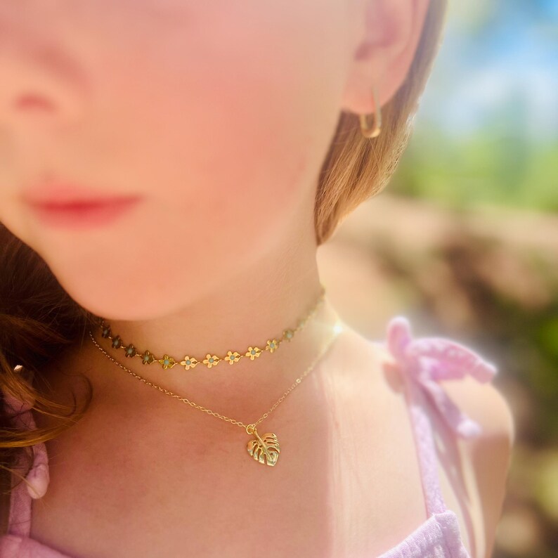 May include: A close-up of a young person wearing a gold necklace with a small leaf pendant and a gold choker necklace with small flower charms. The person is wearing a light purple shirt.