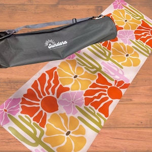 Puede incluir: Una esterilla de yoga rectangular con un patrón repetitivo de flores naranjas, amarillas y rosas con cactus verdes sobre un fondo blanco, acompañada de una bolsa de transporte cilíndrica gris oscuro con el texto blanco Sundara impreso al frente.