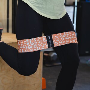 May include: An orange and white leopard print resistance band is wrapped around the upper thigh of a person wearing black leggings. The band has the text "SIMPLY APPLIED" on a black label. The person is in a squat position.