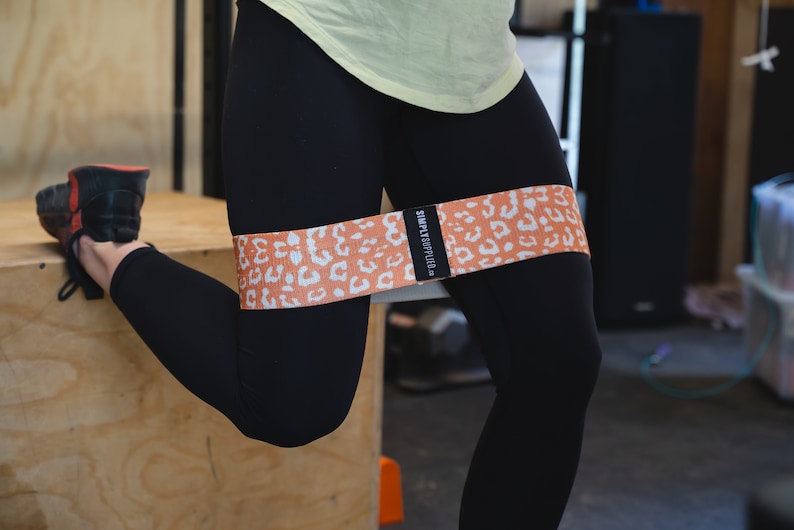 May include: An orange and white leopard print resistance band is wrapped around the upper thigh of a person wearing black leggings. The band has the text "SIMPLY APPLIED" on a black label. The person is in a squat position.
