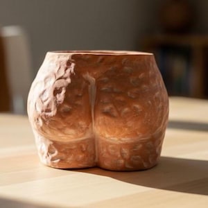 May include: A terracotta-coloured ceramic pot shaped like a human posterior. The pot has a textured surface with detailed sculpting, and is sitting on a wooden table. The pot is designed to be a decorative item.