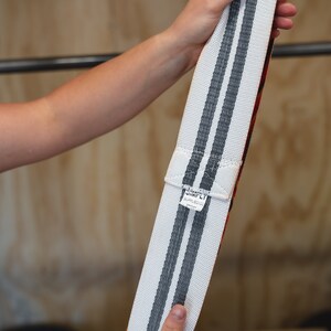 May include: Weightlifting belt with white and gray stripes. The belt has a red interior and a white label with black text. The belt is held up against a wooden background.