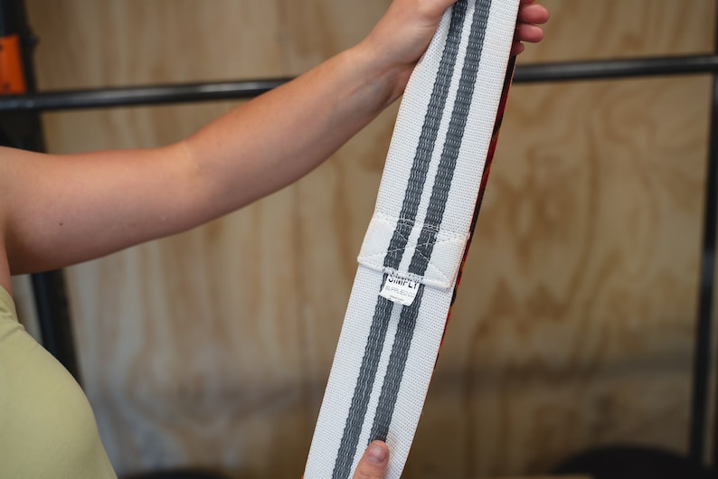 May include: Weightlifting belt with white and gray stripes. The belt has a red interior and a white label with black text. The belt is held up against a wooden background.