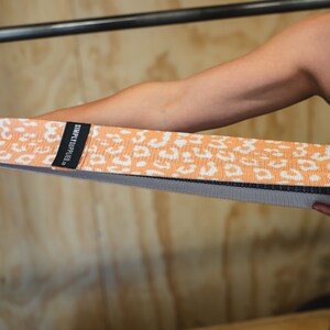 May include: An orange and white patterned resistance band with the text "SIMPLYFITD." The band is held by a person. The band is made of a woven material and has a black border.