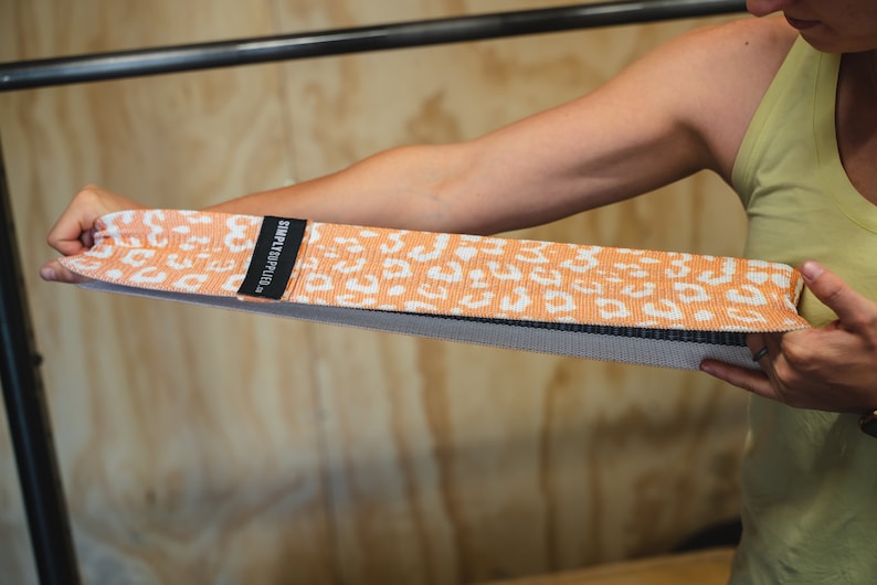 May include: An orange and white patterned resistance band with the text "SIMPLYFITD." The band is held by a person. The band is made of a woven material and has a black border.