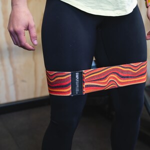 May include: A resistance band with a wavy pattern in shades of red, orange, and black is wrapped around the upper thighs. The band has the text "SIMPLY SUPPLIED" printed on a black label. The person is wearing black leggings.