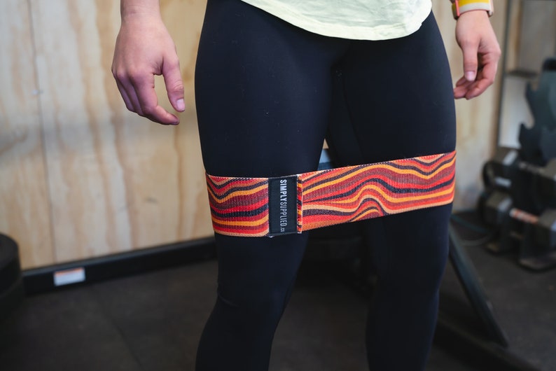 May include: A resistance band with a wavy pattern in shades of red, orange, and black is wrapped around the upper thighs. The band has the text "SIMPLY SUPPLIED" printed on a black label. The person is wearing black leggings.