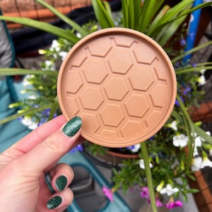 May include: A circular light brown coaster featuring a raised hexagonal honeycomb pattern across its surface. The coaster has a smooth, flat base and a slightly elevated outer rim, providing a structured design for holding a beverage glass.