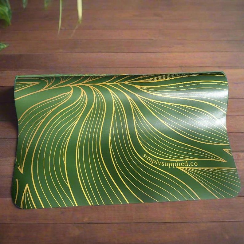 May include: A green yoga mat with a gold leaf design. The mat has a repeating pattern of gold lines that resemble leaves. The text "simplysupplied.co" is printed in white on the mat.