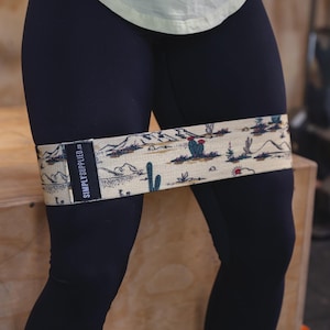 May include: A beige resistance band with a desert landscape pattern, including cacti, mountains, and a setting sun. The band is wrapped around the upper thigh. The text "SIMPLY SUPPLIED.CO" is printed on a black label.