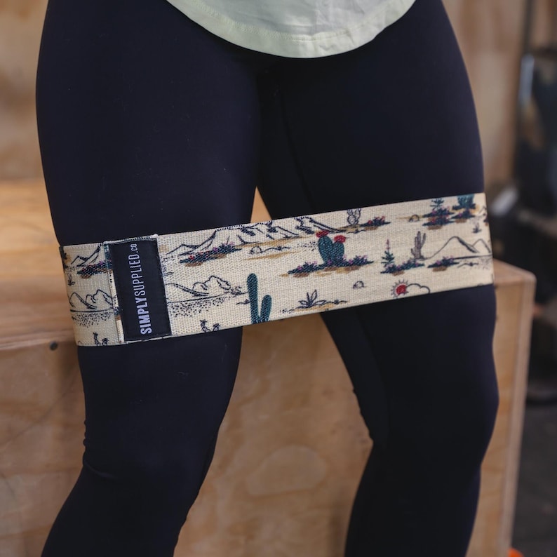 May include: A beige resistance band with a desert landscape pattern, including cacti, mountains, and a setting sun. The band is wrapped around the upper thigh. The text "SIMPLY SUPPLIED.CO" is printed on a black label.