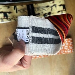 May include: A hand holds a white and gray striped resistance band with the word "SIMPLY" on a tag. The band has a woven texture and is partially folded, revealing a patterned interior with red, orange, and brown stripes. Other bands with different patterns are visible in the background.