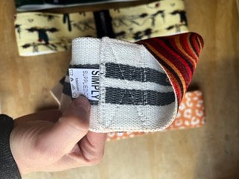May include: A hand holds a white and gray striped resistance band with the word "SIMPLY" on a tag. The band has a woven texture and is partially folded, revealing a patterned interior with red, orange, and brown stripes. Other bands with different patterns are visible in the background.