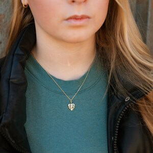 May include: A gold necklace featuring a dainty monstera leaf pendant. The necklace is worn by a person with long blonde hair and a teal blue top.