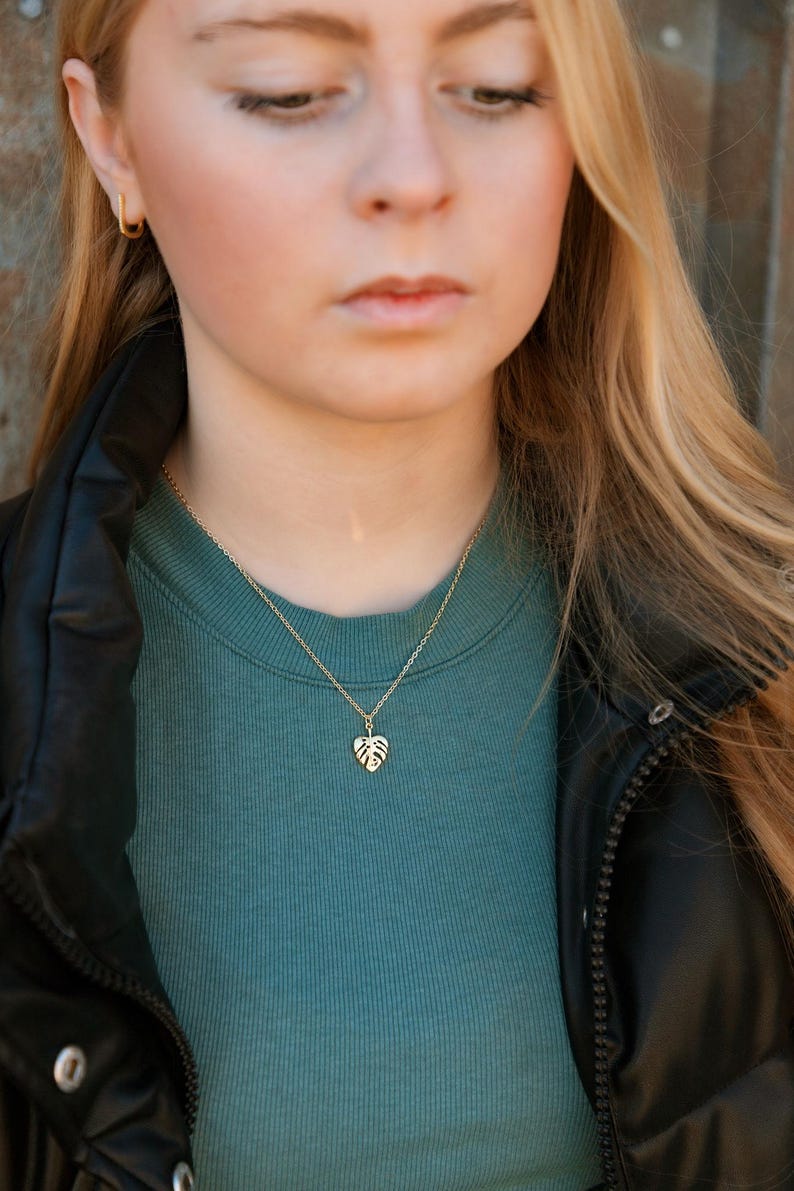 May include: A gold necklace featuring a dainty monstera leaf pendant. The necklace is worn by a person with long blonde hair and a teal blue top.