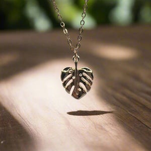 May include: A silver necklace featuring a delicate, cutout monstera leaf pendant. The necklace is hanging from a wooden surface.