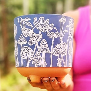 May include: A blue ceramic planter with a white floral and mushroom design. The planter has a terracotta base and three feet.