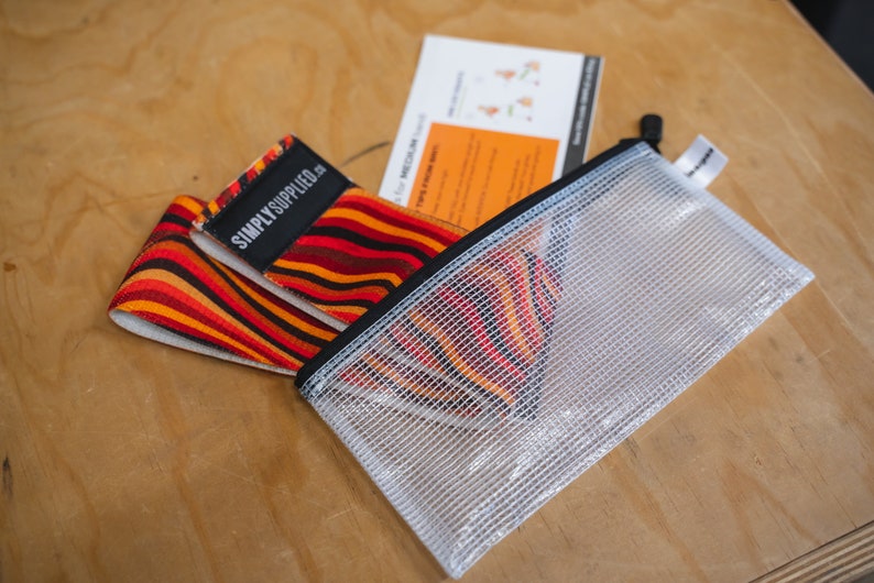 May include: A set of resistance bands with a vibrant orange, red, and brown striped pattern. The bands are labeled "SIMPLY SUPPLIED." They are accompanied by a clear mesh zipper pouch and a small instruction card. The items are on a wooden surface.
