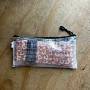 May include: A clear, rectangular zippered pouch with a black zipper and pull. The pouch contains several small, rectangular items with a red and white leopard print design. The pouch is resting on a light brown, wooden surface.