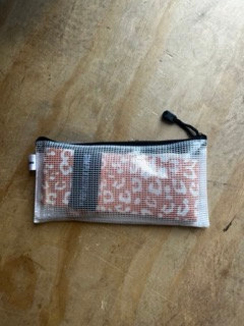 May include: A clear, rectangular zippered pouch with a black zipper and pull. The pouch contains several small, rectangular items with a red and white leopard print design. The pouch is resting on a light brown, wooden surface.
