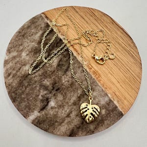 May include: A gold chain necklace with a gold monstera leaf pendant. The necklace is on a wooden and marble surface.