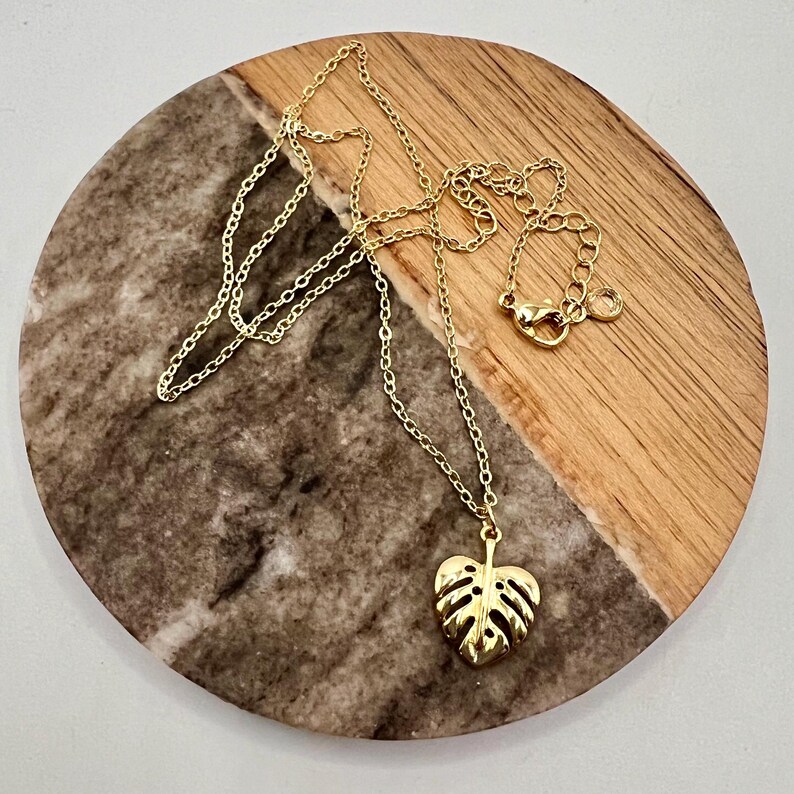 May include: A gold chain necklace with a gold monstera leaf pendant. The necklace is on a wooden and marble surface.