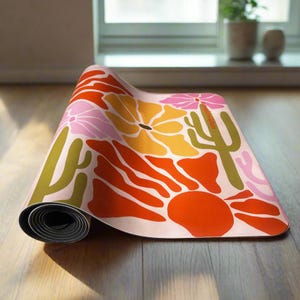 May include: A rolled-up yoga mat with a colorful floral and cactus design. The mat features orange, yellow, pink, and green illustrations on a light pink background. The mat is on a wooden floor.