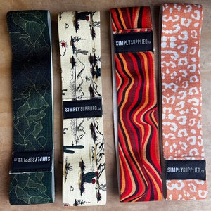 May include: Four resistance bands with unique patterns. One is dark green with gold lines, another has a desert landscape design, a third features red and orange wavy stripes, and the last has a leopard print. Each band has a black label with "SIMPLY SUPPLIED.CO".