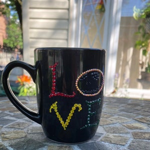 May include: Black ceramic mug with a white 'LOVE' design made of colorful rhinestones. The mug has a handle and is sitting on a stone surface.