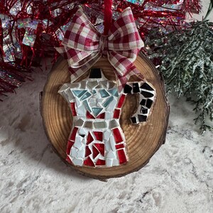 May include: A round wooden slice ornament featuring a mosaic moka pot design made of white, red, light blue, and black glass tiles. The ornament is topped with a red and white checkered fabric bow and hangs from a red string for display.