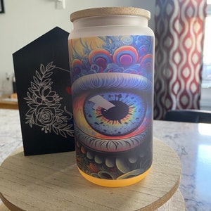 May include: A glass can-shaped tumbler with a bamboo lid. The tumbler features a vibrant, psychedelic eye design with a rainbow iris and colorful background. The base of the tumbler glows with a warm light. A black decorative piece with floral design is behind the tumbler.