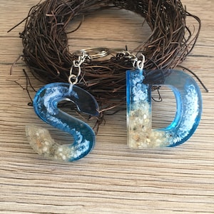 Könnte beinhalten: Ein blauer und weißer Schlüsselanhänger aus Harz mit den Buchstaben "S" und "D", in die Sand eingebettet ist. Der Schlüsselanhänger ist an einer silbernen Kette befestigt.
