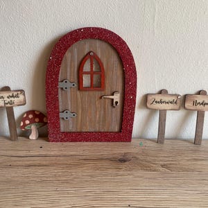 Könnte beinhalten: Eine rote Glitzer-umrahmte Holztür für Feen mit einem kleinen Fenster und einem Türknauf. Es gibt drei Holzschildchen mit dem Text "Hier wohnt Tunte", "Zauberwald" und "Nordpol".