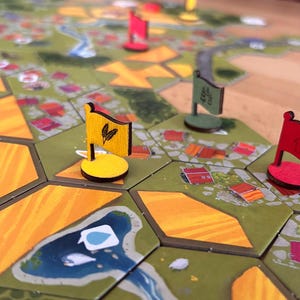 Könnte beinhalten: Ein Brettspiel-Aufbau mit sechseckigen Spielsteinen, die grüne Felder, orangefarbene Ernten und blaue Flüsse zeigen. Mehrere kleine hölzerne Flaggen-Spielsteine in Gelb, Rot und Grün stehen auf den Feldern, wobei die gelbe Flagge ein schwarzes Weizensymbol trägt.