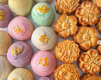 May include: Assortment of colorful mooncakes with various designs and patterns. The mooncakes are arranged in two rows on a white surface. Some mooncakes are decorated with sesame seeds.