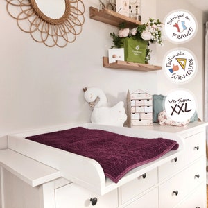 Peut inclure: Une table à langer blanche avec un matelas à langer texturé violet foncé. La table à langer fait partie d'une commode blanche avec des boutons noirs. Un jouet en peluche en forme de cygne blanc est à proximité.
