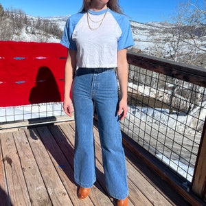 May include: A woman wearing a white and blue baseball tee shirt with a blue and white striped pattern on the sleeves. She is wearing high-waisted, wide-leg blue jeans.