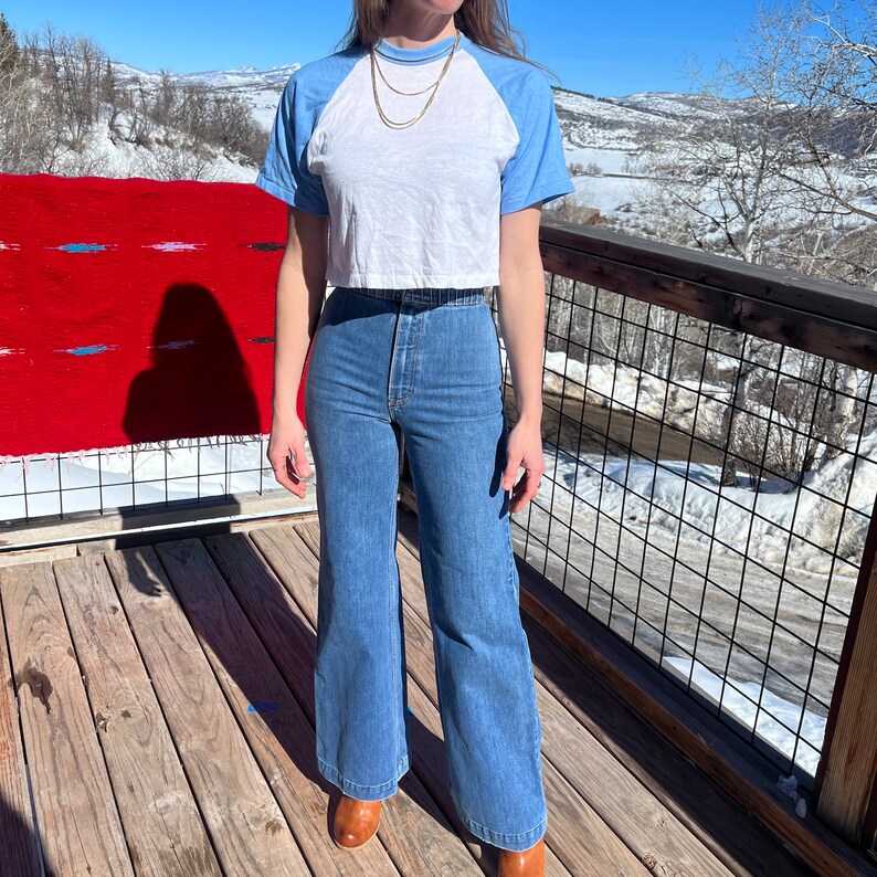 May include: A woman wearing a white and blue baseball tee shirt with a blue and white striped pattern on the sleeves. She is wearing high-waisted, wide-leg blue jeans.