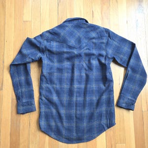 May include: A blue plaid long-sleeve shirt with a button-down front and a pointed collar. The shirt features a classic plaid pattern in shades of blue and tan. The back of the shirt has a yoke detail.