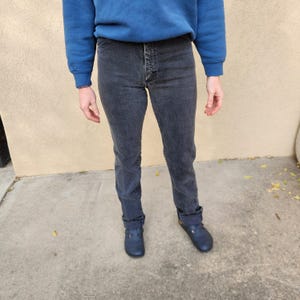 May include: A person wearing a blue sweatshirt and dark gray denim jeans. The jeans have a button fly and are cuffed at the ankles. The person is also wearing blue shoes.