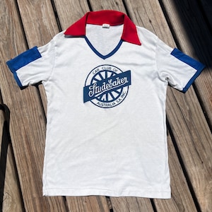 May include: White vintage t-shirt with a red collar and blue sleeve accents. The shirt features a blue logo that reads "Car Club of Studebaker Australia S.A. Inc." in a circular design. The shirt has a V-neck.