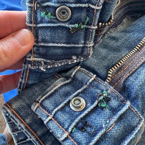 May include: Close-up of a blue denim jacket with a zipper and two button closures. The buttons are metal and have a silver finish. The jacket has a distressed look with faded areas and visible stitching.