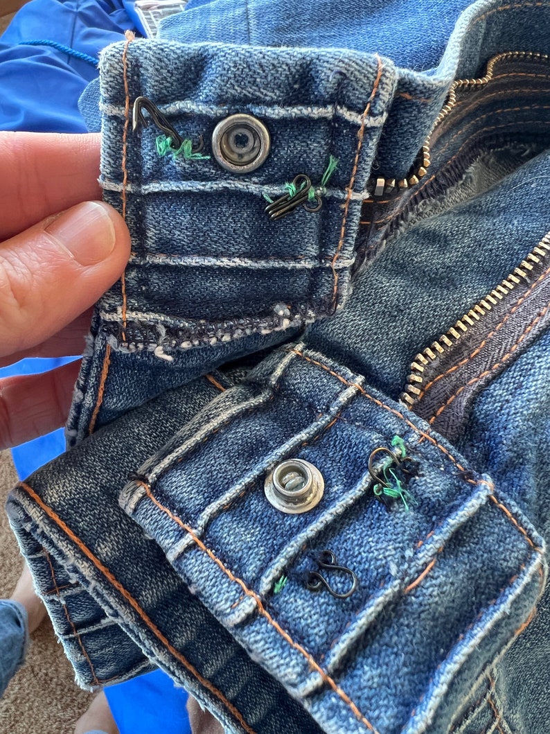 May include: Close-up of a blue denim jacket with a zipper and two button closures. The buttons are metal and have a silver finish. The jacket has a distressed look with faded areas and visible stitching.