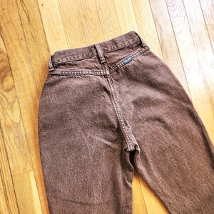Pode incluir: Um par de jeans Wrangler castanhos, vistos por tr&aacute;s. Os jeans t&ecirc;m cintura alta e um design cl&aacute;ssico de cinco bolsos. O logotipo da Wrangler &eacute; vis&iacute;vel em um pequeno patch. O tecido tem uma apar&ecirc;ncia ligeiramente desbotada.