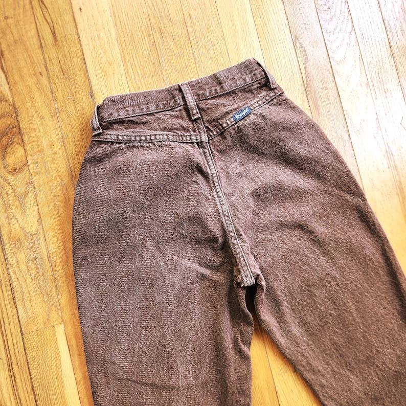 Pode incluir: Um par de jeans Wrangler castanhos, vistos por tr&aacute;s. Os jeans t&ecirc;m cintura alta e um design cl&aacute;ssico de cinco bolsos. O logotipo da Wrangler &eacute; vis&iacute;vel em um pequeno patch. O tecido tem uma apar&ecirc;ncia ligeiramente desbotada.
