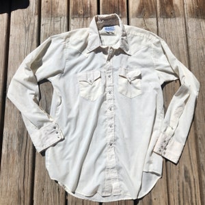 May include: Cream-colored long-sleeve shirt with a Western-style design. Features two chest pockets, pointed collar, and snap closures. The shirt is made of a lightweight fabric and has a classic, vintage aesthetic.