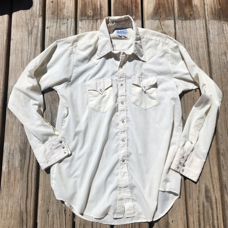 May include: Cream-colored long-sleeve shirt with a Western-style design. Features two chest pockets, pointed collar, and snap closures. The shirt is made of a lightweight fabric and has a classic, vintage aesthetic.