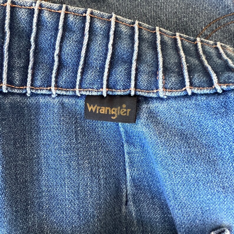 May include: Close-up of a blue denim fabric with a gold Wrangler label sewn on. The fabric has a textured, woven appearance.