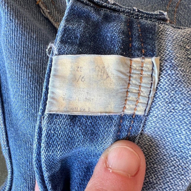 May include: Close-up of a blue denim fabric with a white tag sewn into the fabric. The tag has the word "SIZE" and the number "36" written on it.